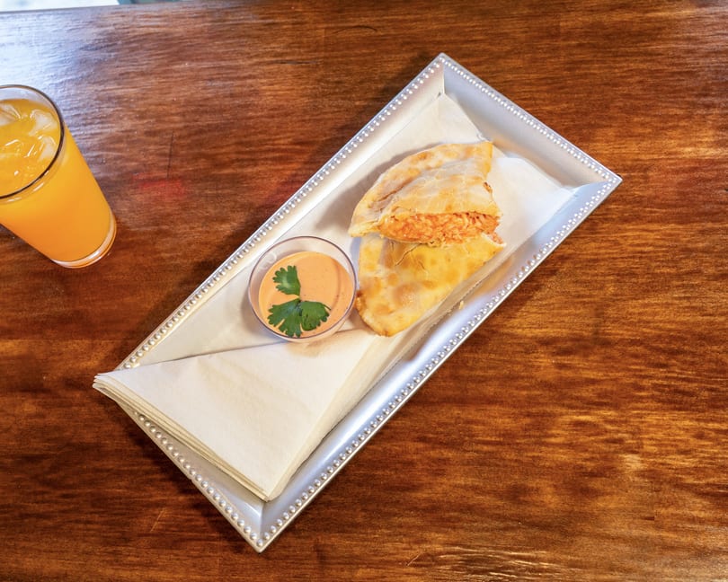 A golden-brown empanada with a creamy dipping sauce and an orange drink on a wooden table.