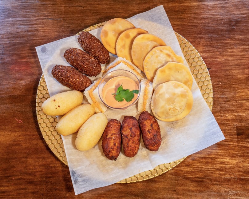 Assortment of fried appetizers including empanadas, croquettes, and corn fritters with dipping sauce.