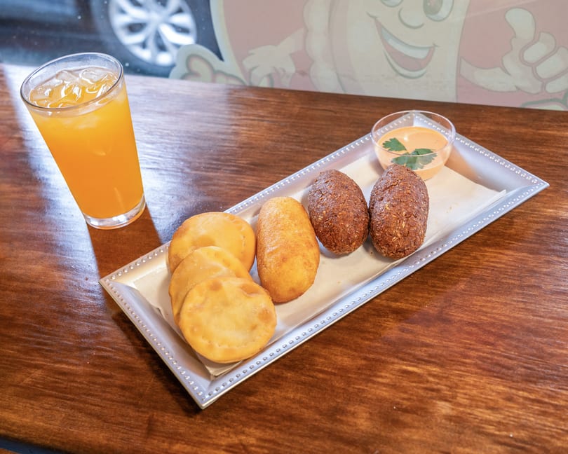 A tray of fried pastries and kibbeh, with a side of sauce and a glass of orange juice.