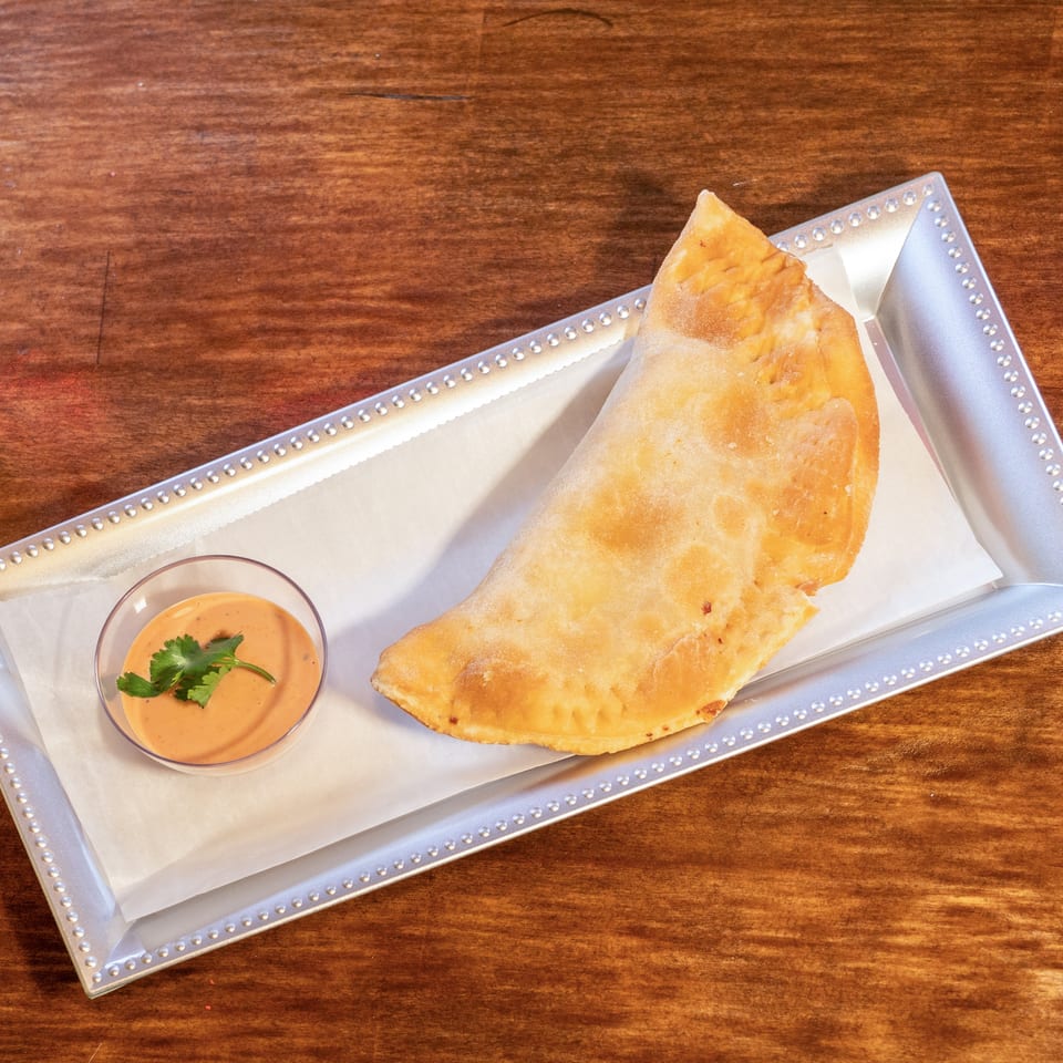 A golden-brown empanada with a side of dipping sauce garnished with cilantro on a silver tray.
