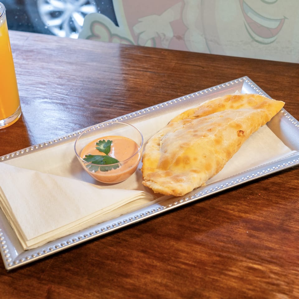 Golden-brown empanada with dipping sauce and orange juice on a wooden table.