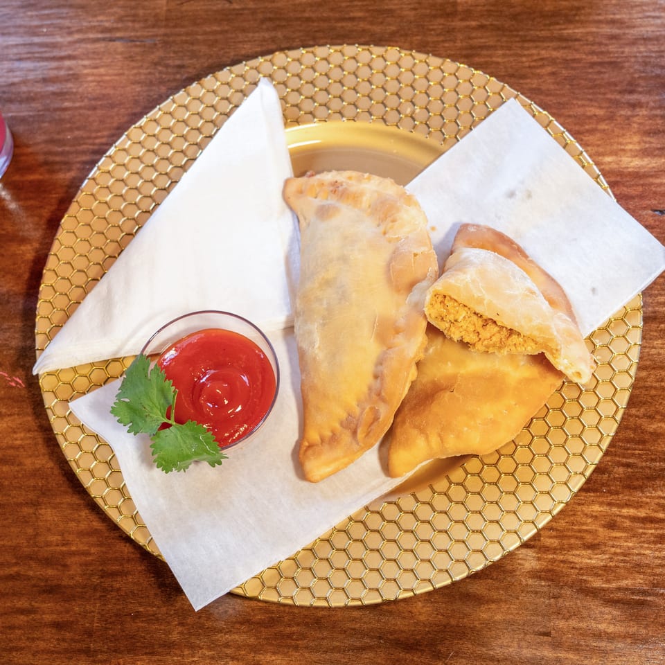 Three golden-brown empanadas on a gold honeycomb plate with ketchup and cilantro, next to a pink drink.