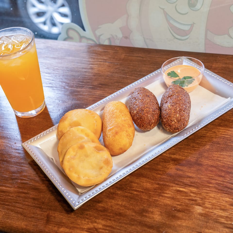A silver tray holds fried pastries, croquettes, and a dipping sauce, with a glass of orange juice.