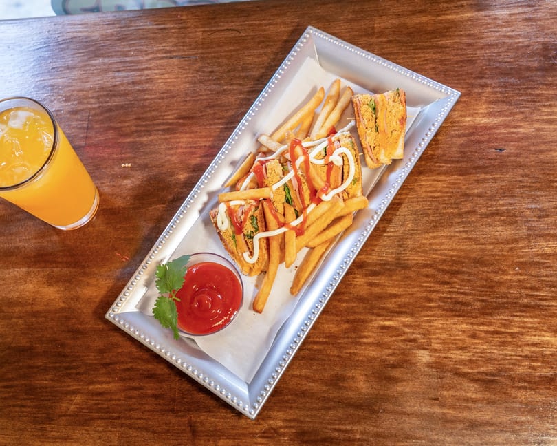 A silver tray holds a club sandwich, fries with ketchup and mayonnaise, and a side of ketchup, next to an orange juice.