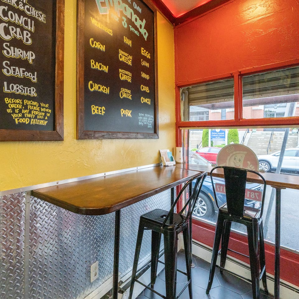 A small restaurant interior with a bar-height table, two stools, and menu boards on the wall.