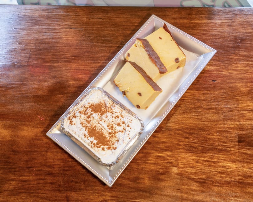 Three slices of bread pudding with raisins and a container of tiramisu on a silver tray.
