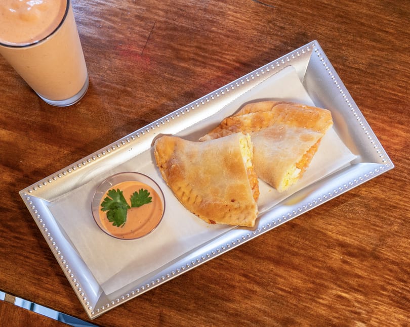 Two golden-brown empanadas with a side of dipping sauce and a smoothie on a wooden table.