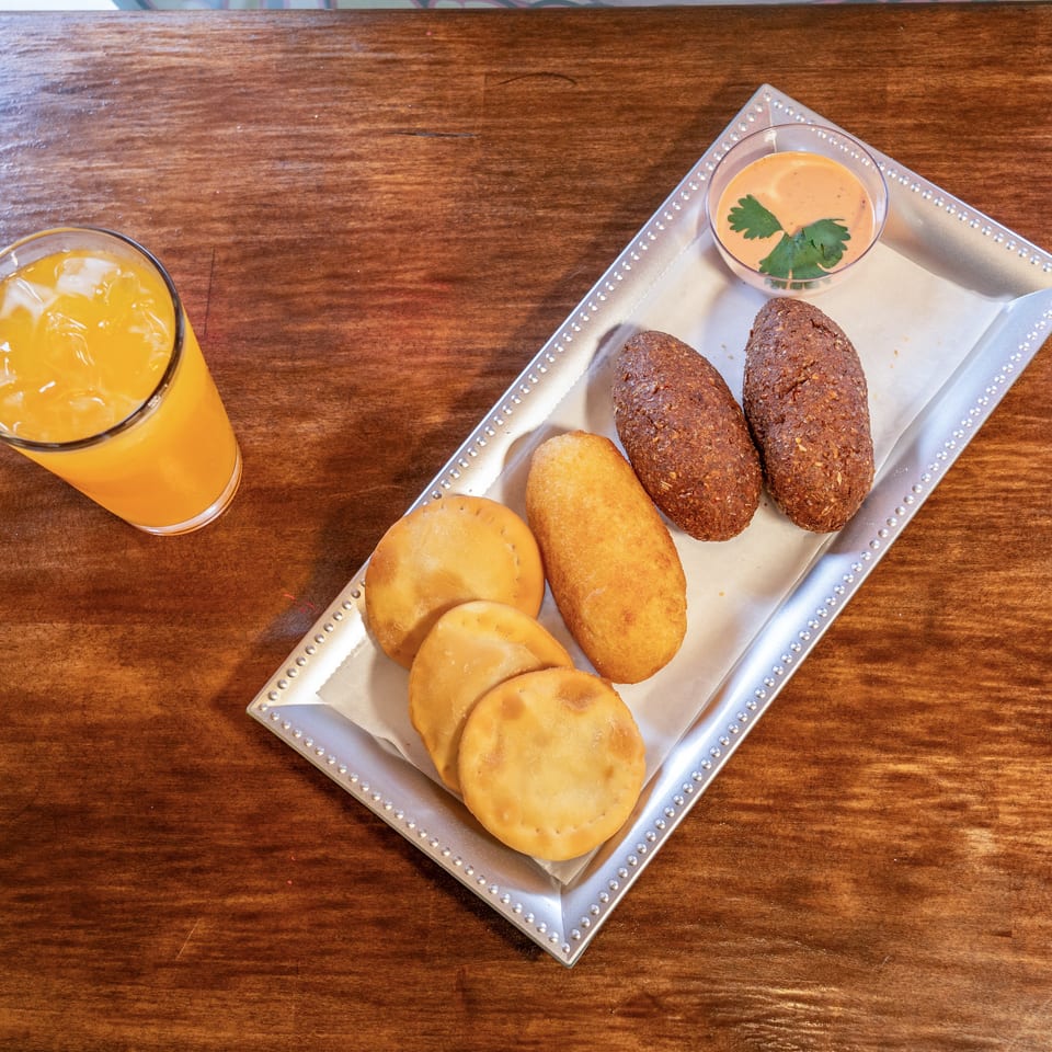 A silver tray with various fried pastries and a dipping sauce, next to a glass of orange juice.