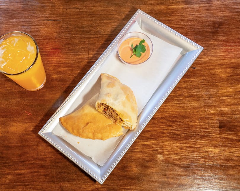 Two empanadas, one cut open to show a savory filling, with dipping sauce and orange juice.