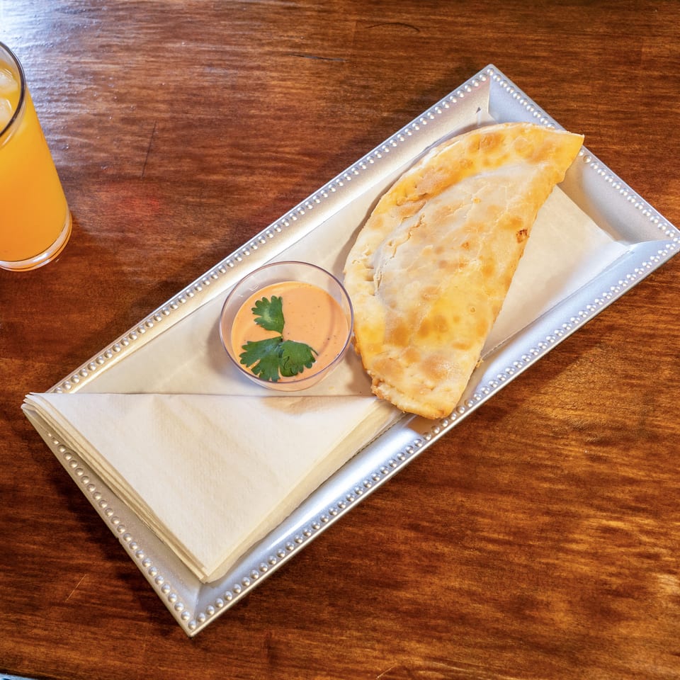 A golden-brown empanada with dipping sauce and an orange drink on a wooden table.