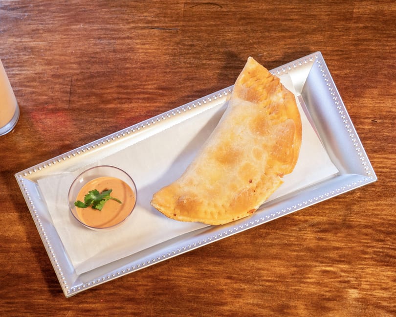 A golden-brown empanada and a small dish of dipping sauce with cilantro on a silver tray.