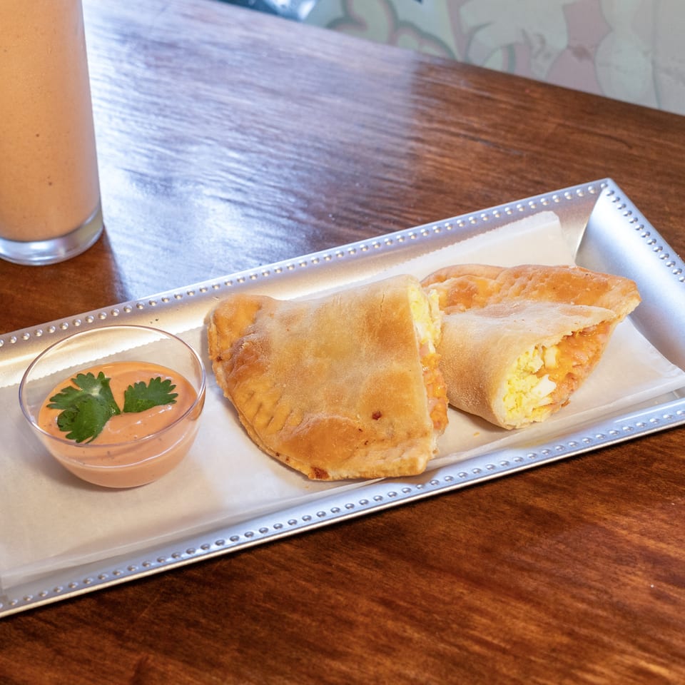 Two golden-brown empanadas, one cut open to show egg and cheese filling, with a side of dipping sauce.