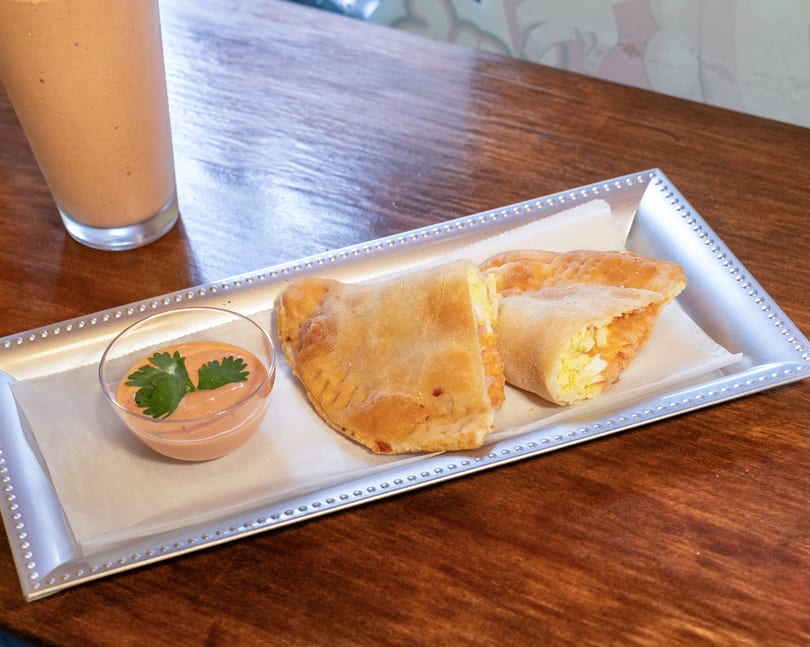 Two golden-brown empanadas, one cut open to show egg and cheese filling, with a side of dipping sauce.